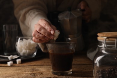 Photo of Brewing aromatic coffee in moka pot. Woman putting sugar cubes in glass with drink at wooden table, closeup