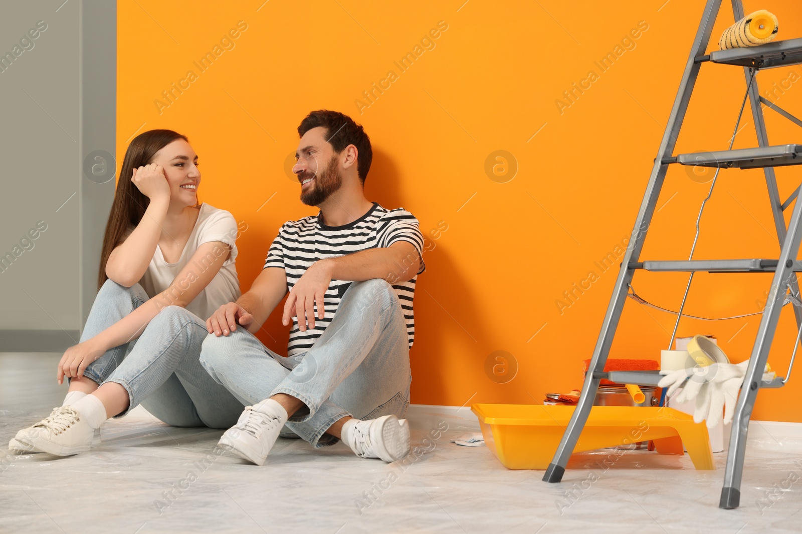 Photo of Happy designers sitting on floor with painting equipment near freshly painted orange wall indoors