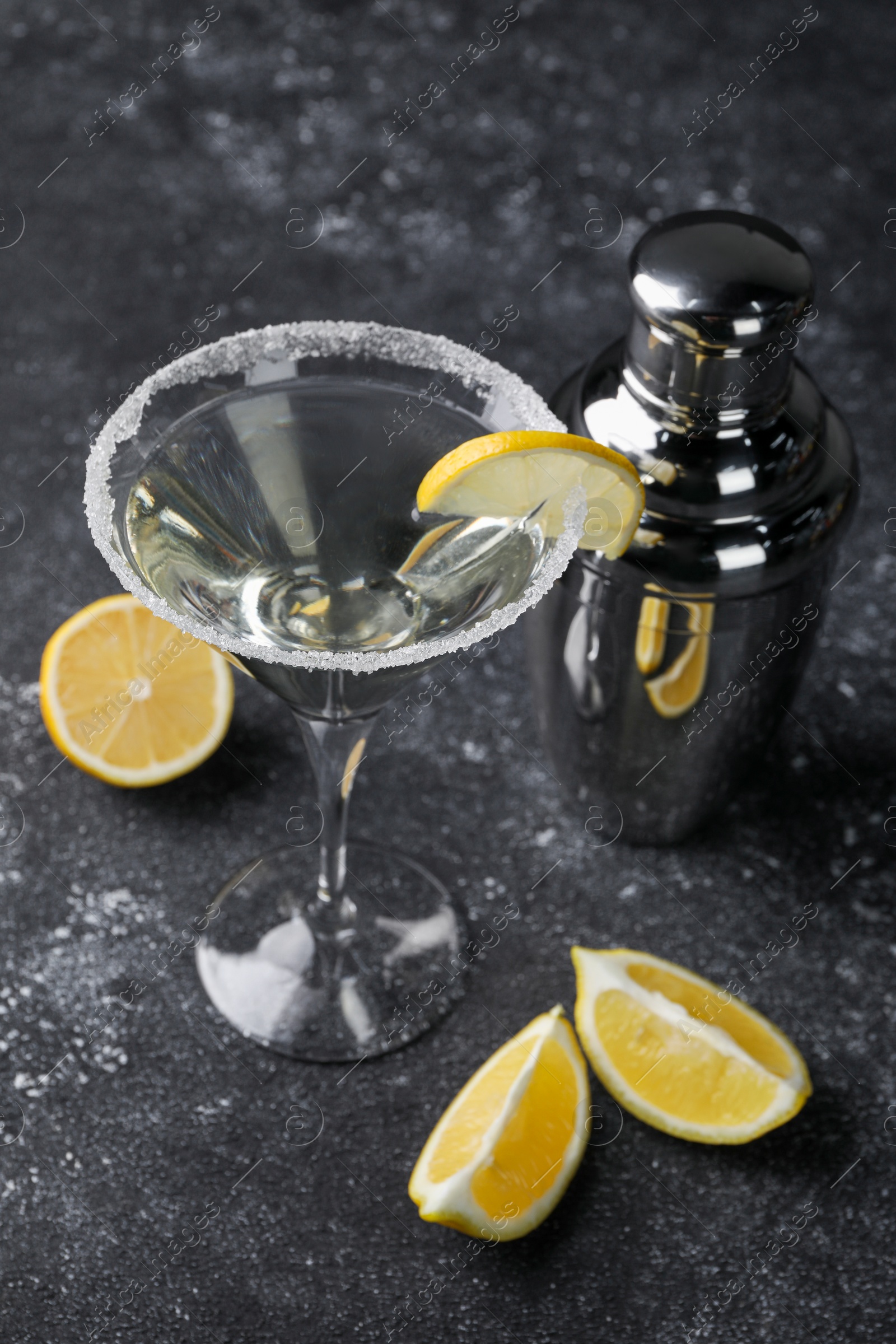 Photo of Martini glass of refreshing cocktail decorated with lemon slice and sugar near shaker on black textured table