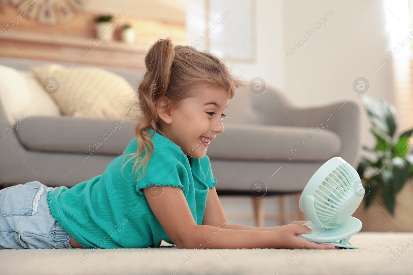 Photo of Little girl enjoying air flow from portable fan on floor in living room. Summer heat