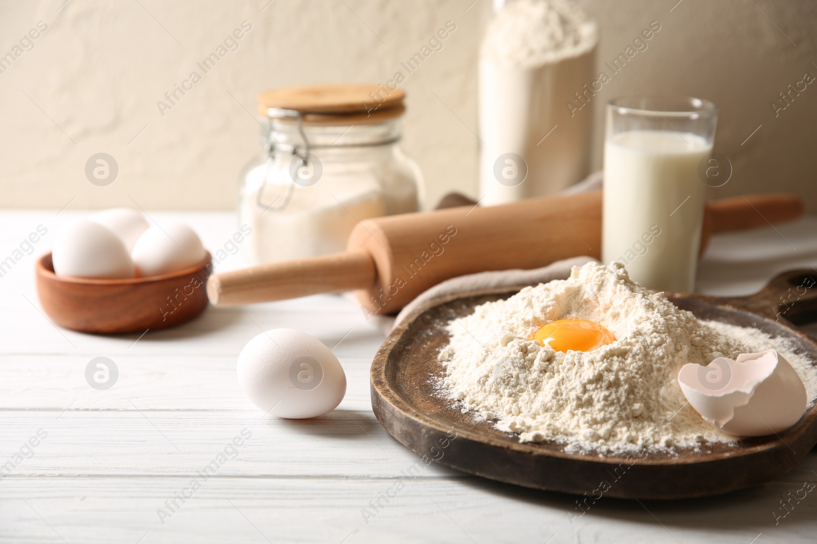 Photo of Making dough. Pile of flour with yolk, rolling pin and eggs on white wooden table, closeup