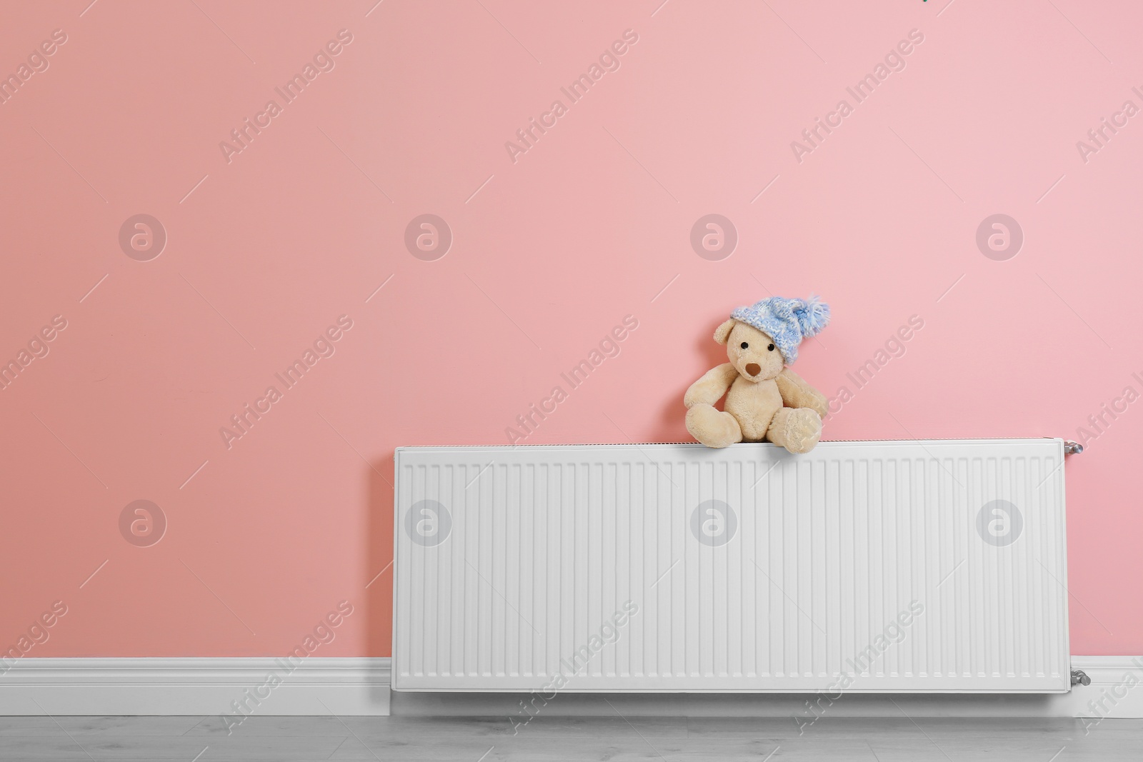 Photo of Teddy bear with knitted hat on heating radiator near color wall. Space for text