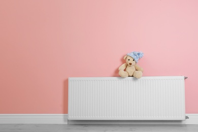 Teddy bear with knitted hat on heating radiator near color wall. Space for text