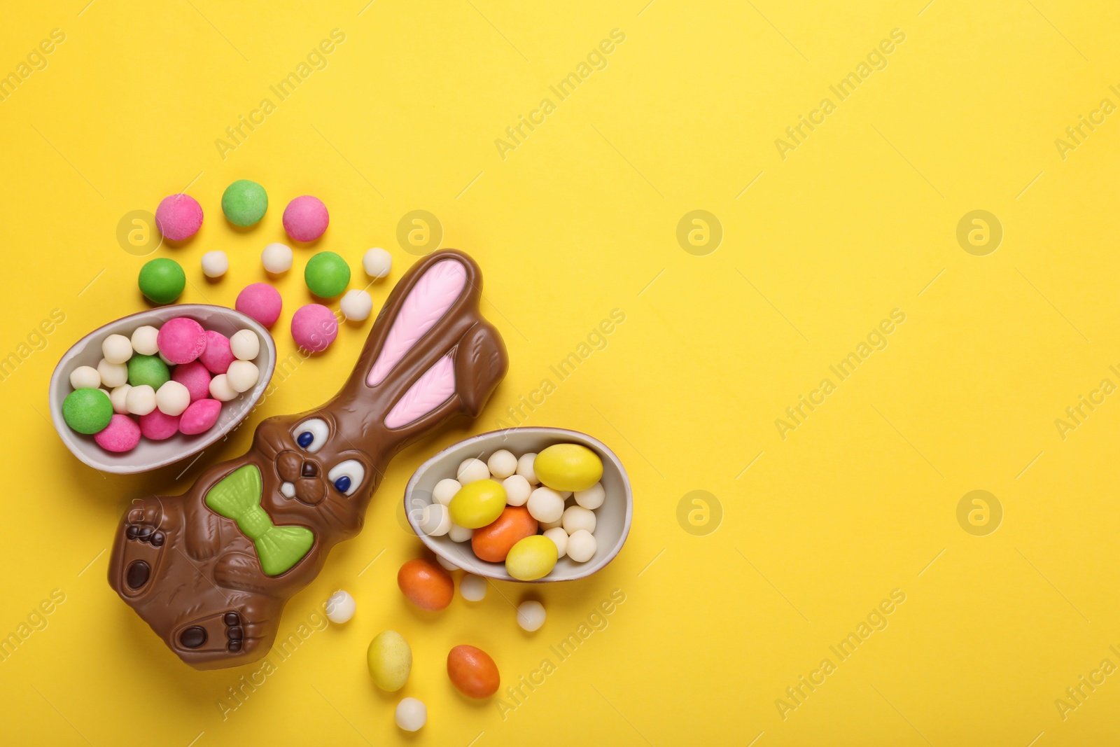 Photo of Chocolate Easter bunny, halves of egg and candies on yellow background, flat lay. Space for text