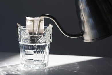 Glass with drip coffee bag and metal kettle on light grey table, closeup