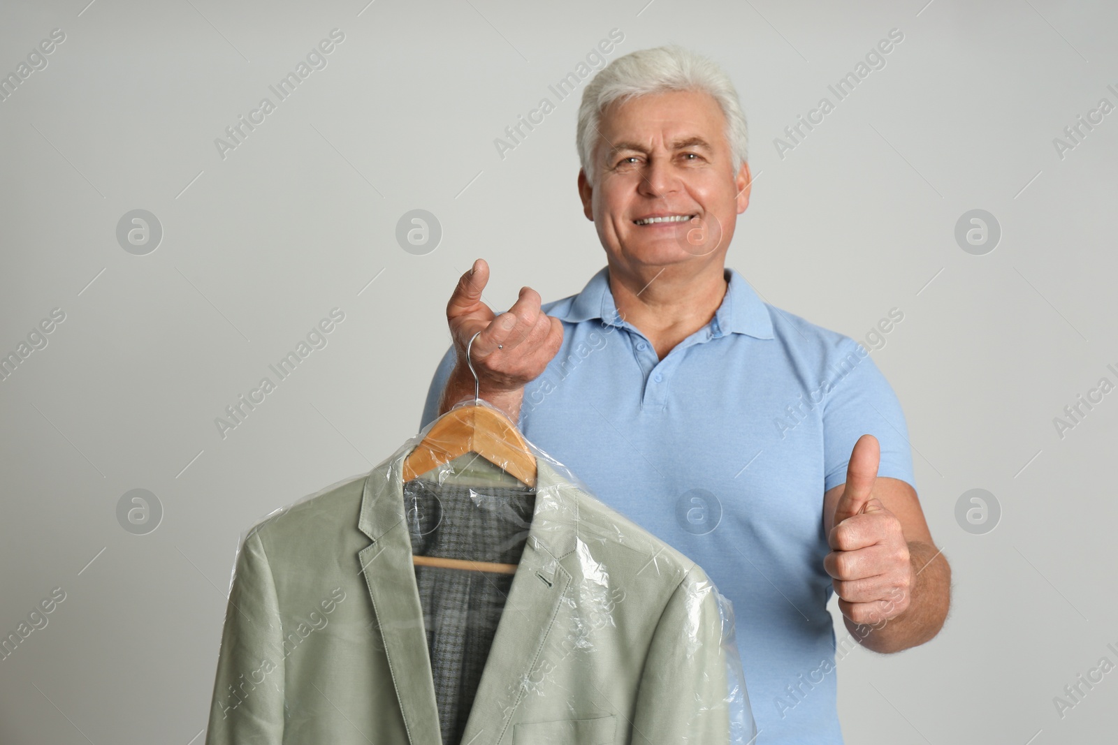 Photo of Senior man holding hanger with jacket in plastic bag on light grey background. Dry-cleaning service