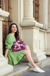 Beautiful woman with bouquet of spring flowers near building outdoors, space for text