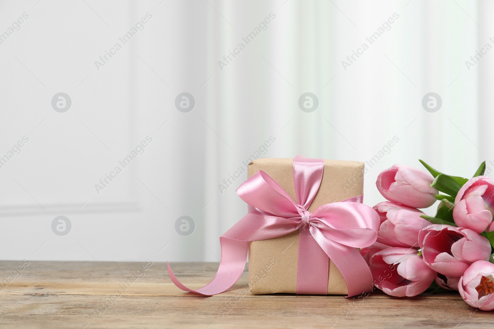 Photo of Beautiful gift box with bow and pink tulips on wooden table. Space for text