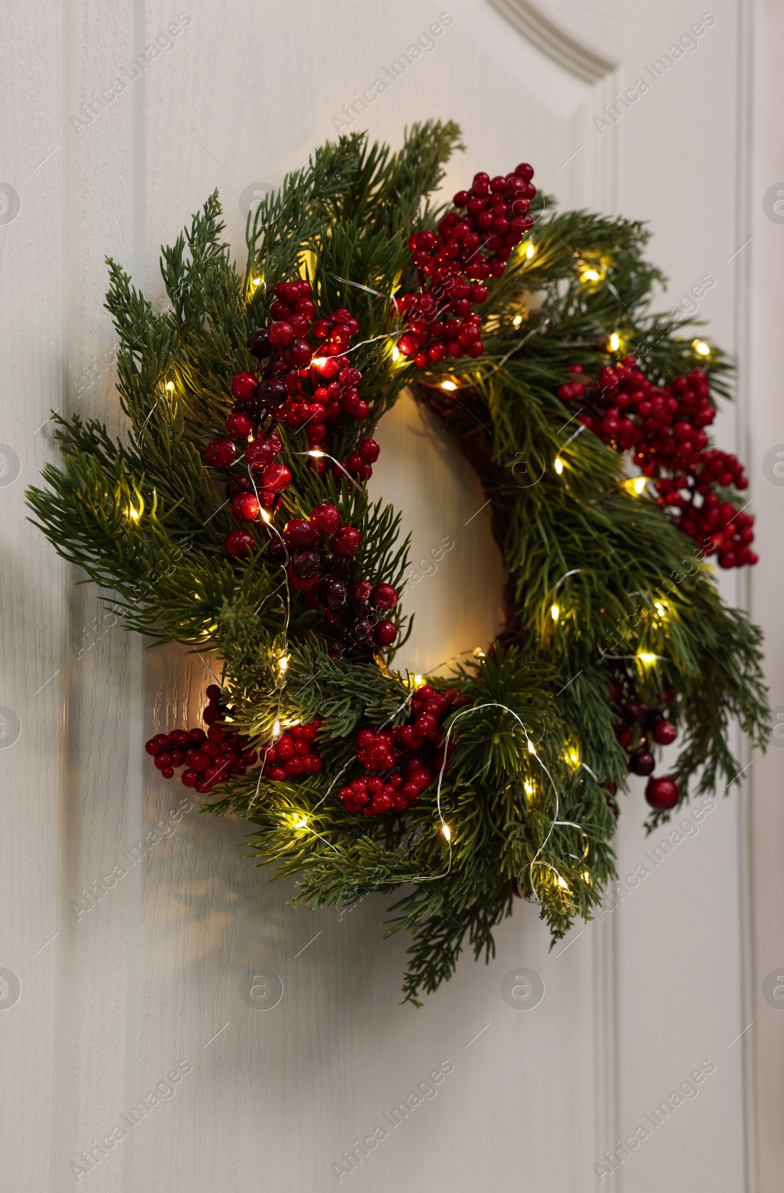 Photo of Beautiful Christmas wreath with red berries and fairy lights hanging on white door