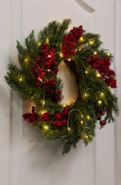 Beautiful Christmas wreath with red berries and fairy lights hanging on white door