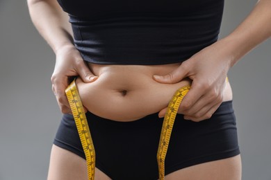 Woman with measuring tape touching belly fat on grey background, closeup. Overweight problem