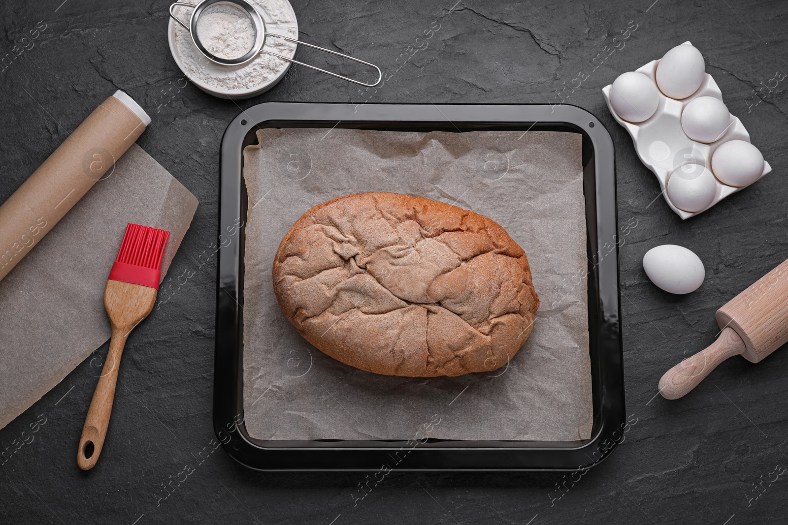 Photo of Baking pan with bread, parchment paper and ingredients on black slate background, flat lay