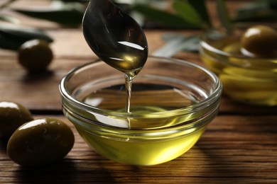 Photo of Spoon with cooking oil over bowl and olives on wooden table, closeup