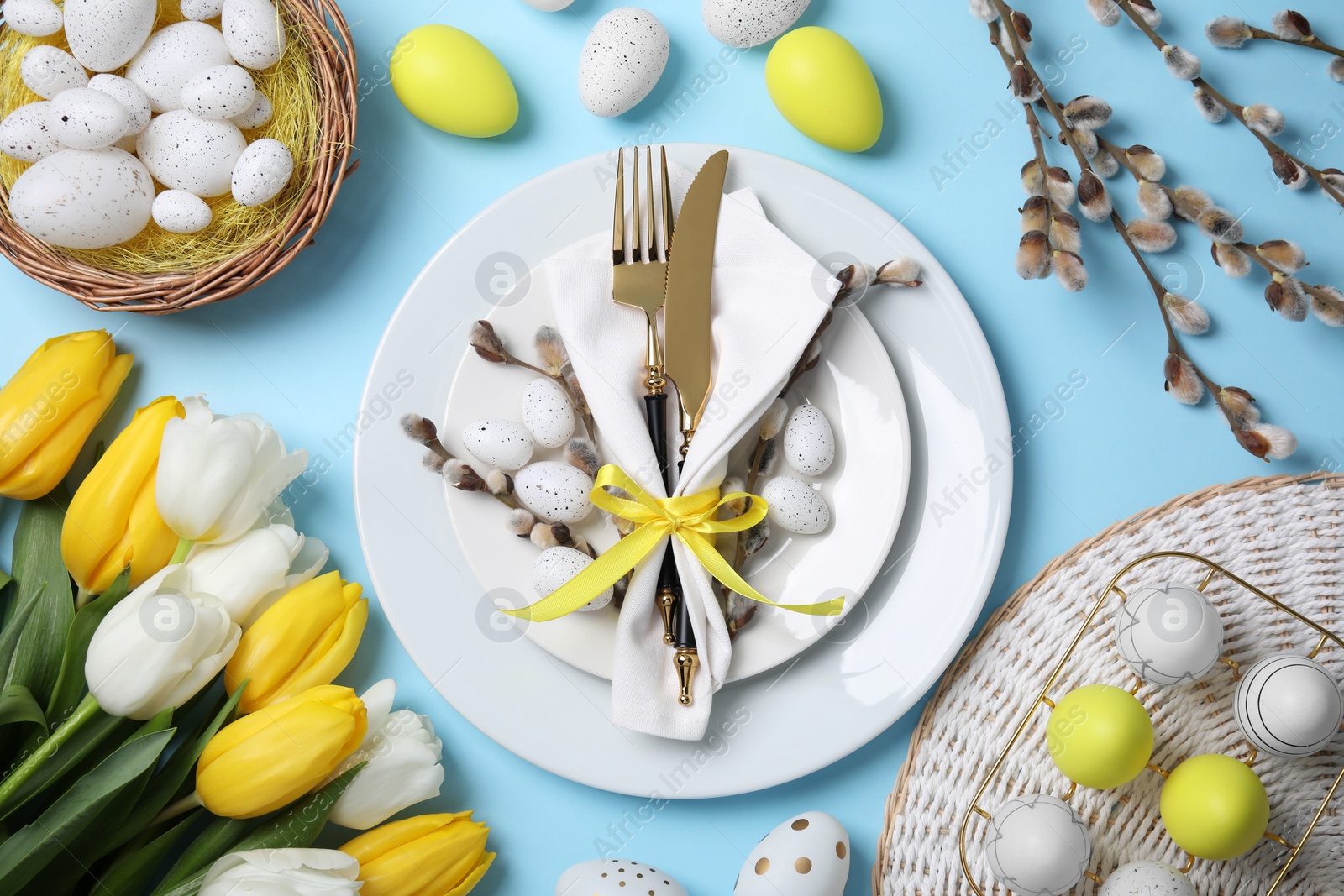 Photo of Festive table setting with tulips and painted eggs on light blue background, flat lay. Easter celebration