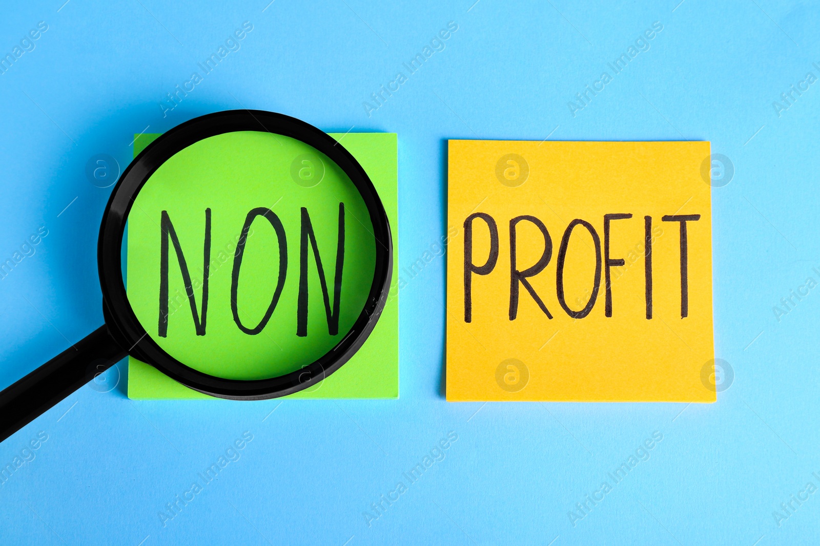 Photo of Paper notes with phrase Non Profit under magnifying glass on light blue background, flat lay