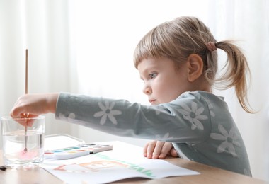 Photo of Cute little girl drawing with brush at wooden table indoors. Child`s art