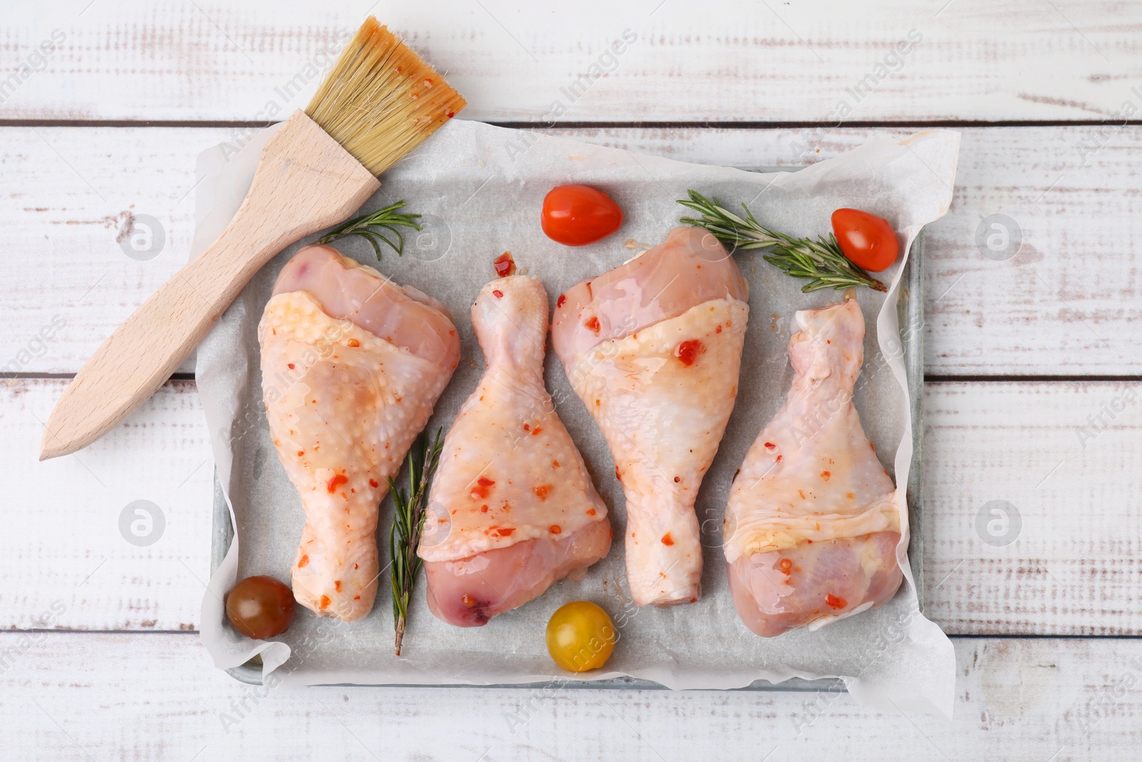 Photo of Raw marinated chicken drumsticks, rosemary, tomatoes and basting brush on white wooden table, flat lay
