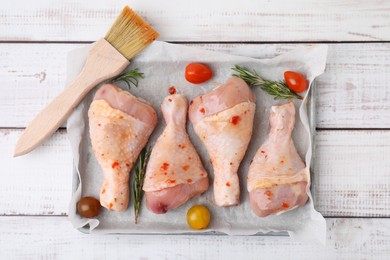 Photo of Raw marinated chicken drumsticks, rosemary, tomatoes and basting brush on white wooden table, flat lay