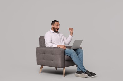 Photo of Happy young man with laptop sitting in armchair on grey background