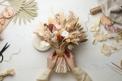 Florist making beautiful bouquet of dried flowers at white table, top view