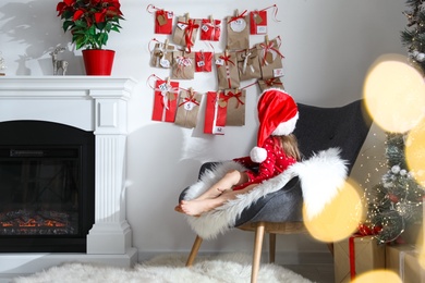 Cute little girl in Santa hat near Christmas advent calendar at home