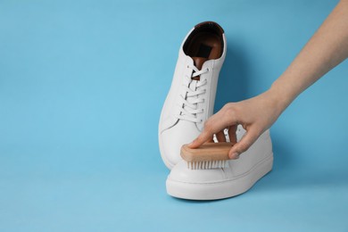 Photo of Woman cleaning stylish white sneakers with brush on light blue background, closeup. Space for text