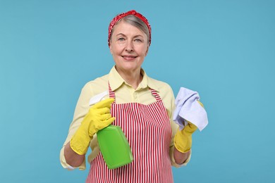 Happy housewife with spray bottle and rag on light blue background
