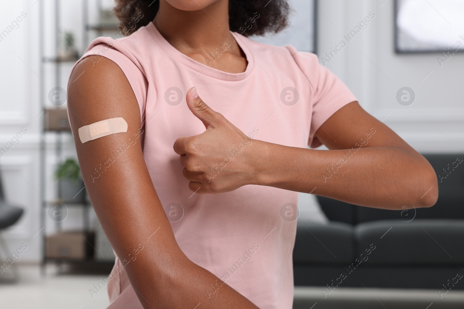 Photo of Young woman with adhesive bandage on her arm after vaccination showing thumb up indoors, closeup