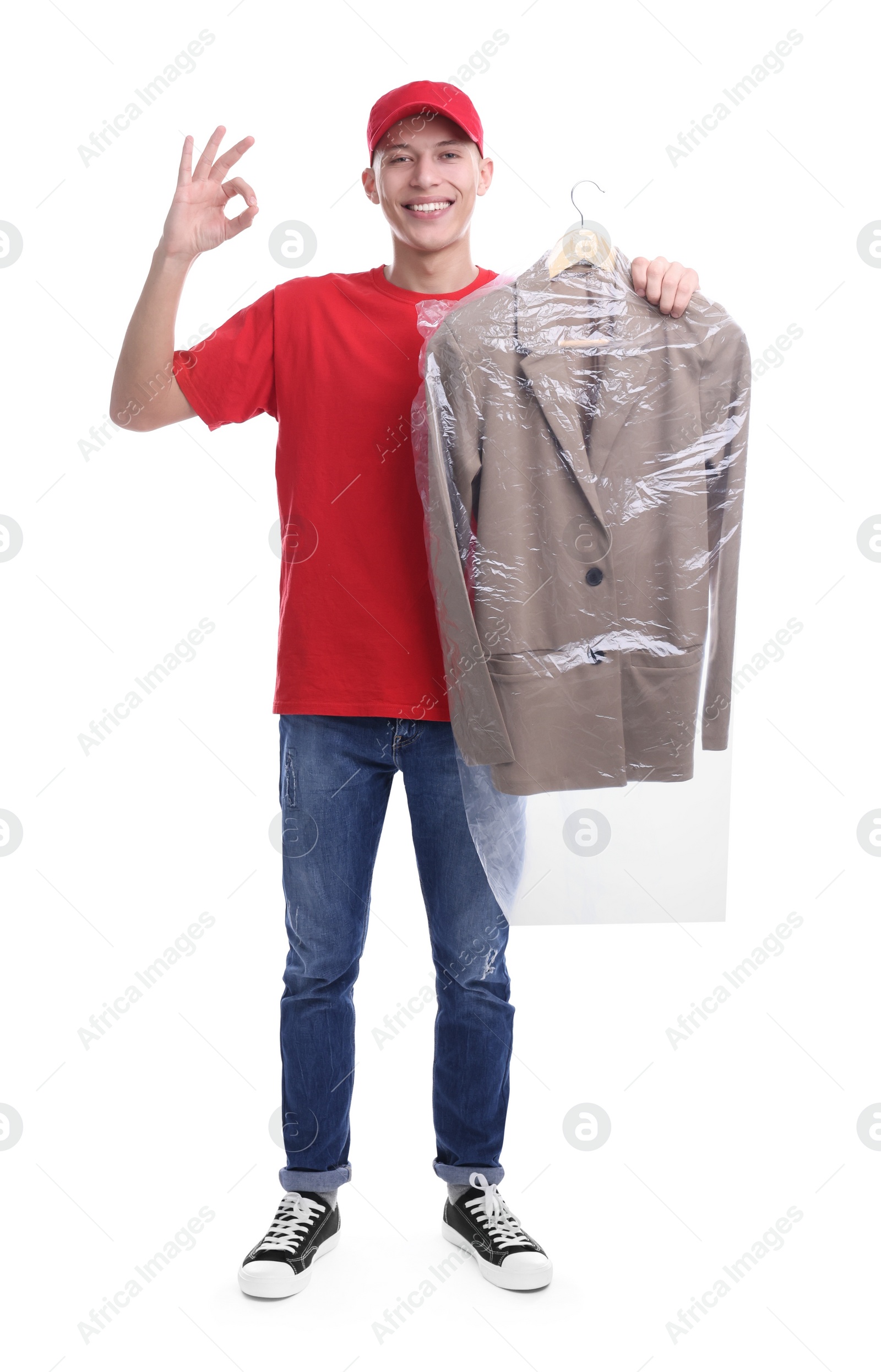 Photo of Dry-cleaning delivery. Happy courier holding jacket in plastic bag and showing OK gesture on white background