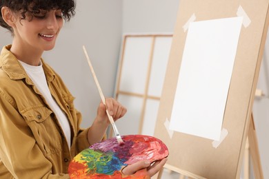 Photo of Young woman mixing paints on palette with brush near easel in studio