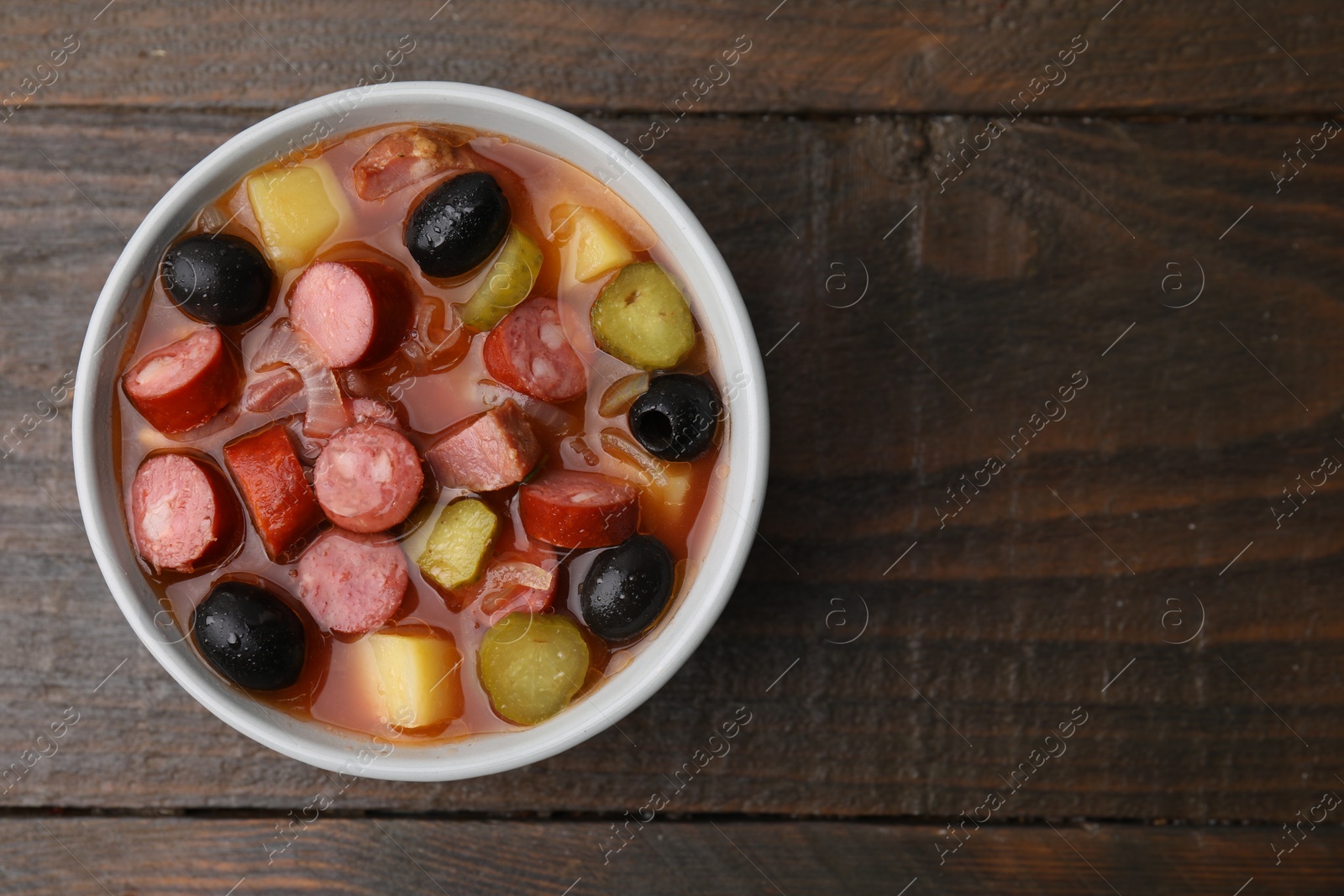 Photo of Meat solyanka soup with thin dry smoked sausages in bowl on wooden table, top view. Space for text