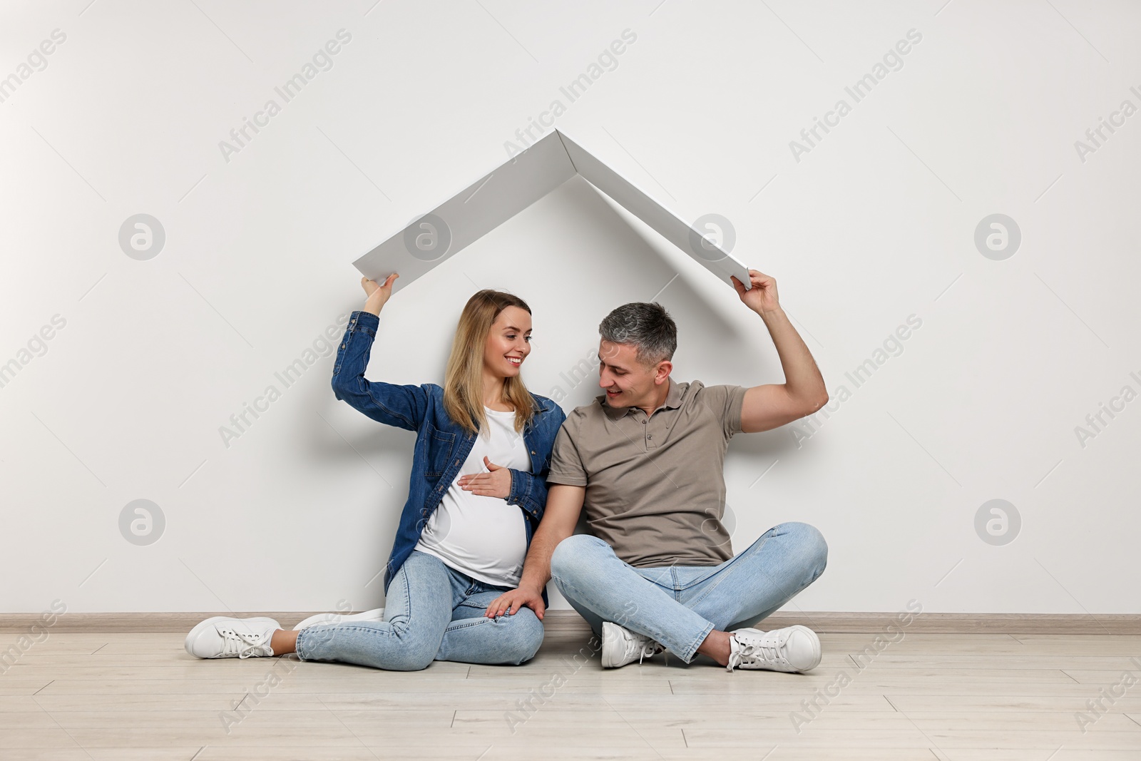 Photo of Young family housing concept. Pregnant woman with her husband sitting under cardboard roof on floor indoors