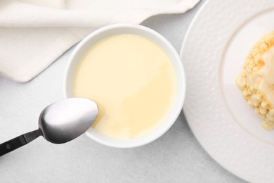 Photo of Tasty condensed milk in bowl and spoon on light grey table, flat lay