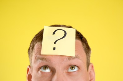 Emotional man with question mark on yellow  background, closeup