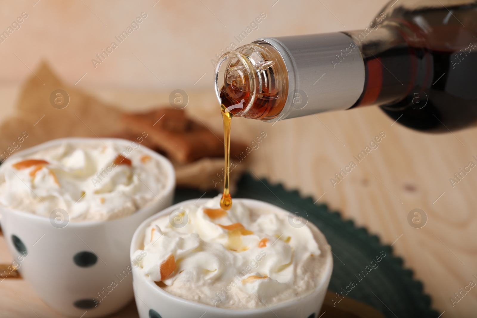 Photo of Pouring delicious caramel syrup into cup with coffee and whipped cream at wooden table, closeup
