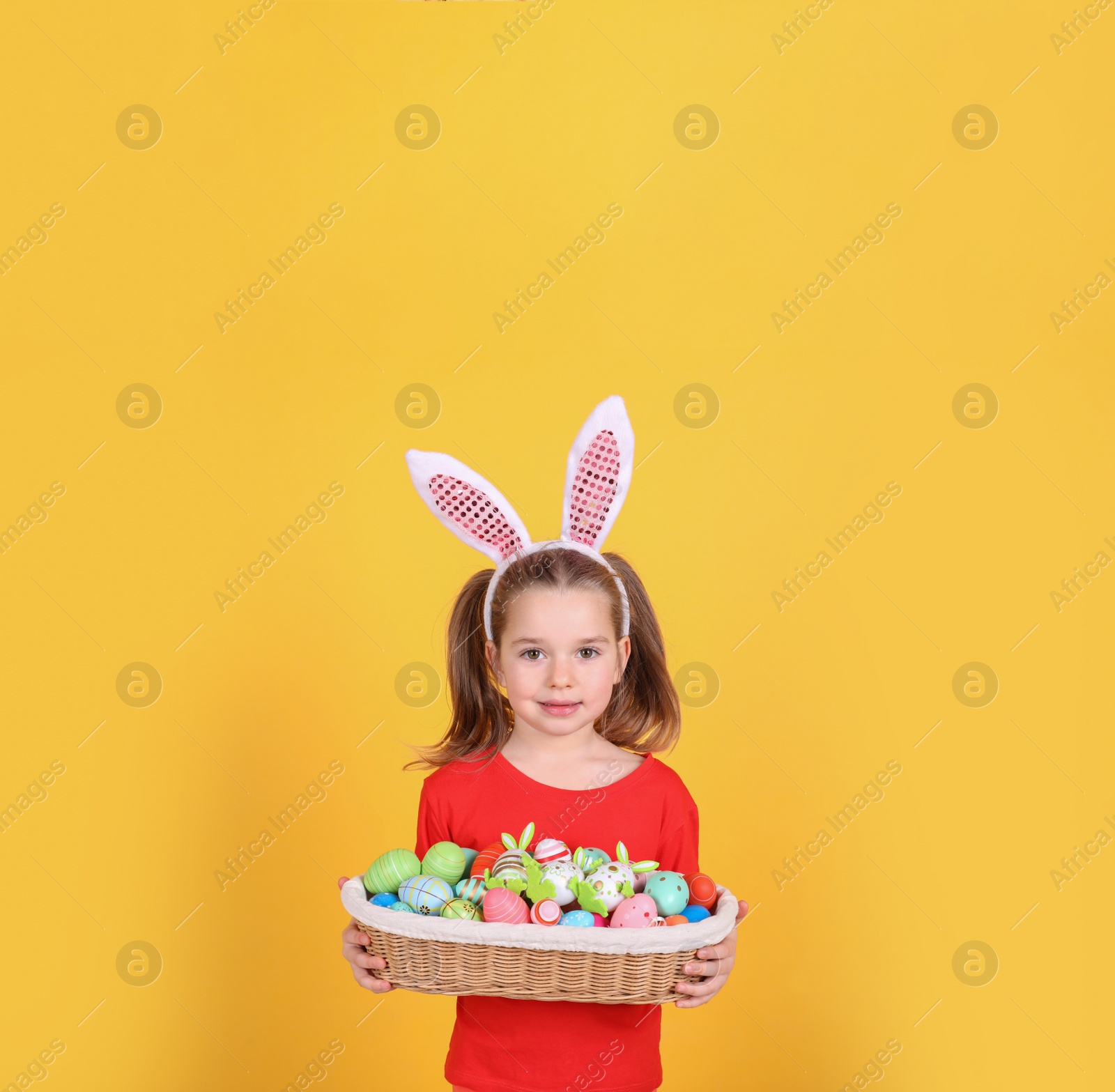 Photo of Adorable little girl with bunny ears holding wicker basket full of Easter eggs on orange background