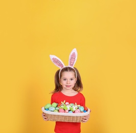Adorable little girl with bunny ears holding wicker basket full of Easter eggs on orange background