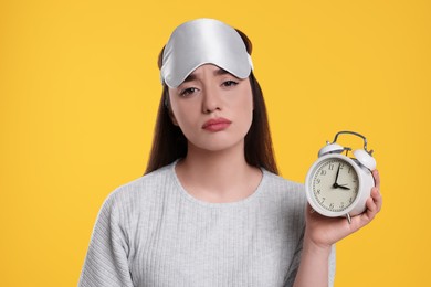 Photo of Tired young woman with sleep mask and alarm clock on yellow background. Insomnia problem
