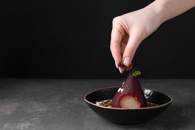 Woman putting muesli on tasty red wine poached pear in bowl at grey table, closeup. Space for text