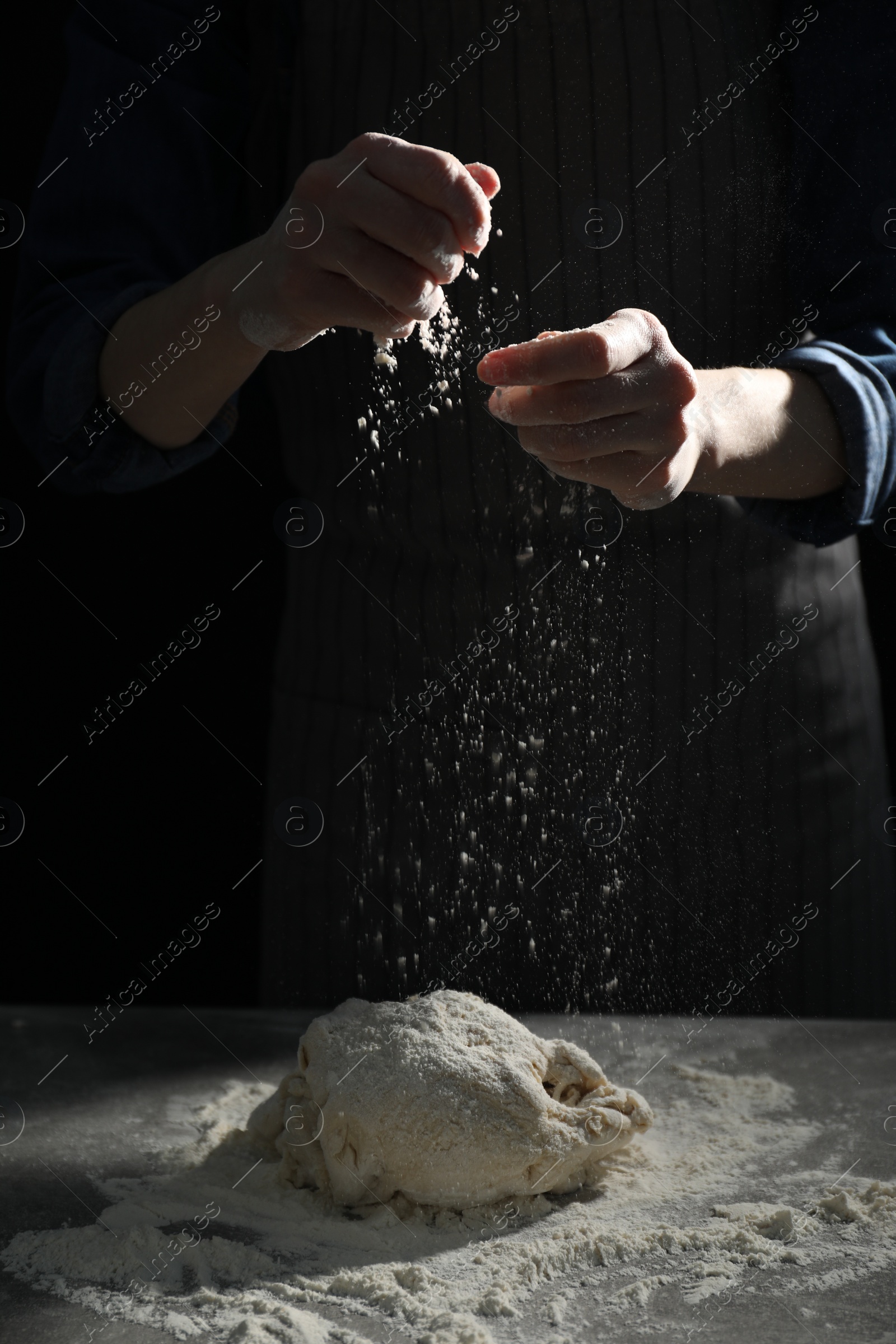 Photo of Making bread. Woman sprinkling flour over dough at table on dark background, closeup