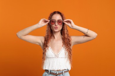 Photo of Stylish young hippie woman in sunglasses sending air kiss on orange background