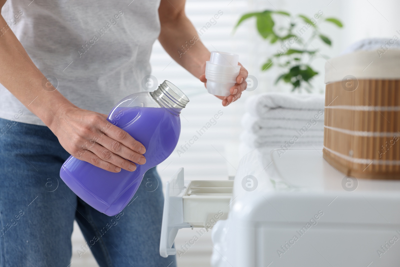 Photo of Woman pouring fabric softener from bottle into cap near washing machine in bathroom, closeup