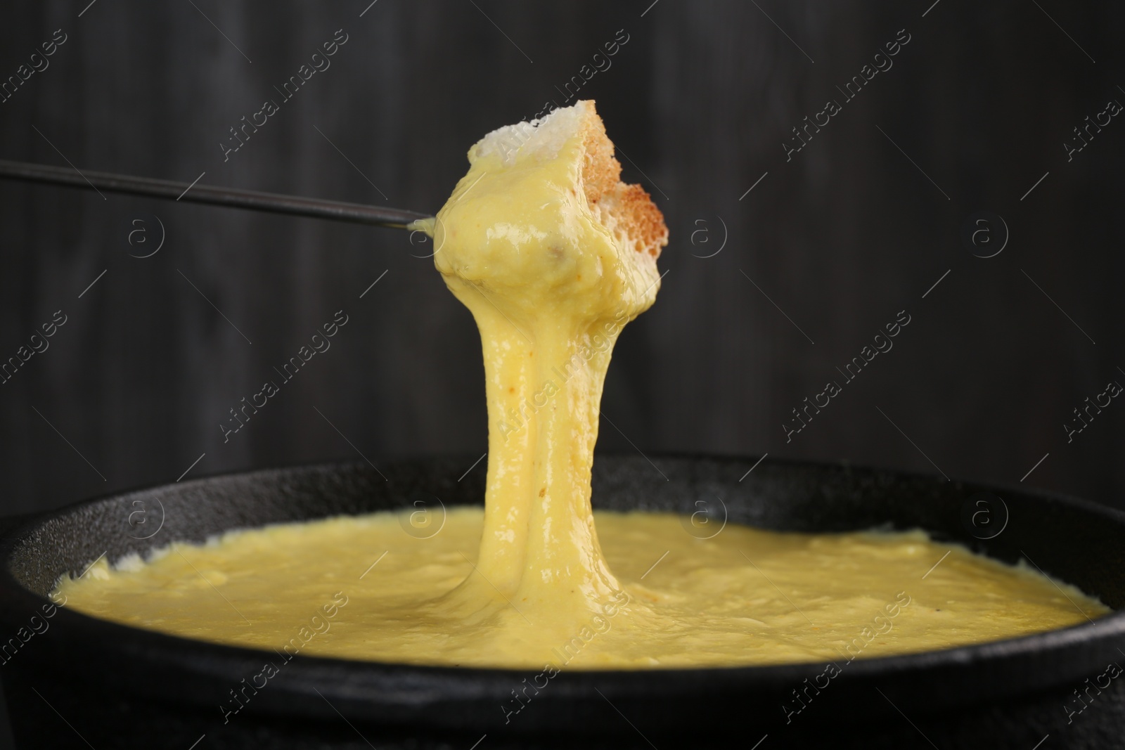 Photo of Dipping piece of bread into fondue pot with tasty melted cheese against dark gray background, closeup