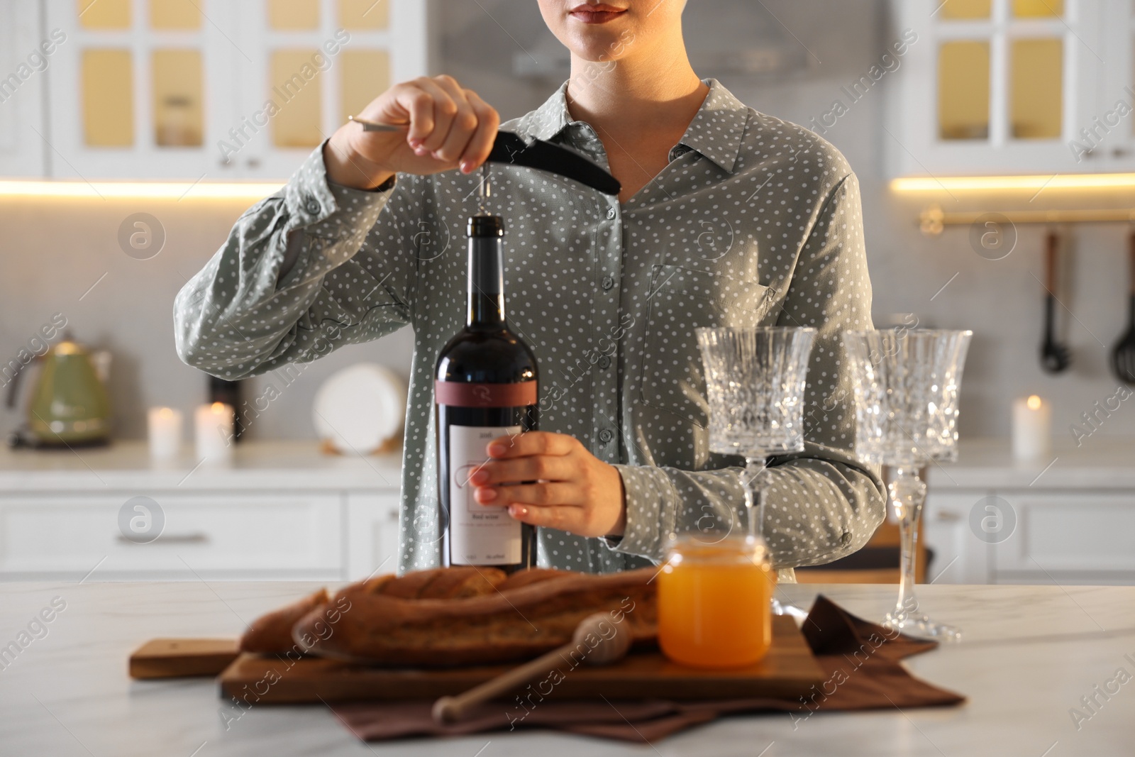 Photo of Romantic dinner. Woman opening wine bottle with corkscrew at table in kitchen, closeup