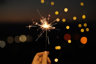 Woman holding bright sparkler against blurred lights, closeup