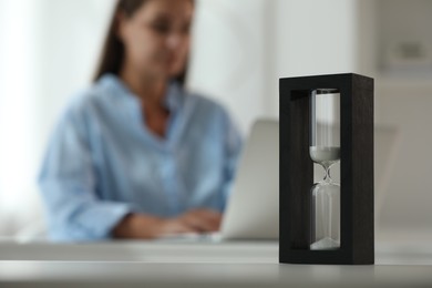 Photo of Hourglass with flowing sand on white table. Woman using laptop indoors, selective focus