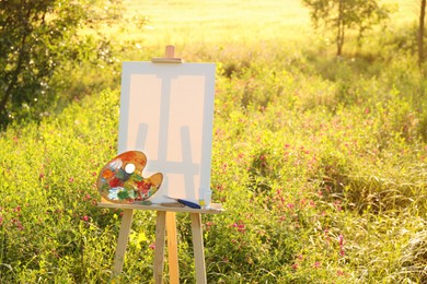 Photo of Wooden easel with blank canvas and painting equipment in meadow. Space for text