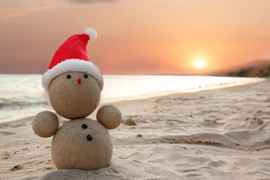 Photo of Snowman made of sand with Santa hat on beach near sea at sunset, space for text. Christmas vacation