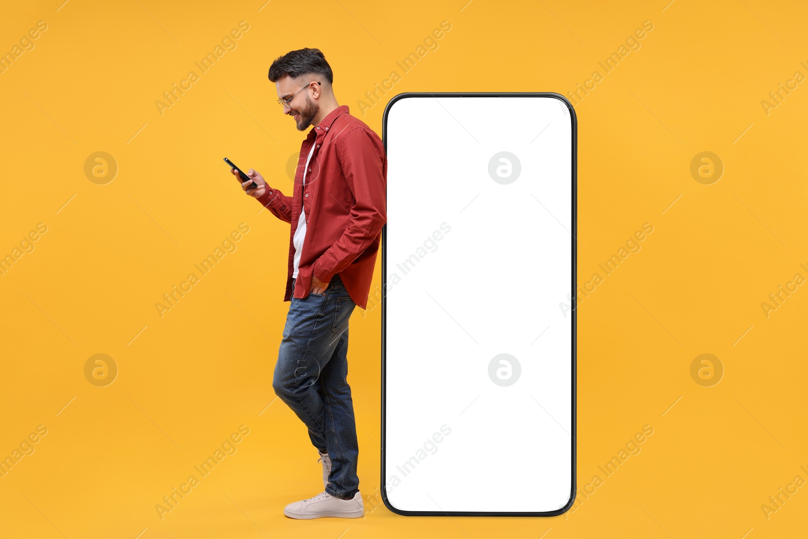 Image of Man with mobile phone standing near huge device with empty screen on orange background. Mockup for design