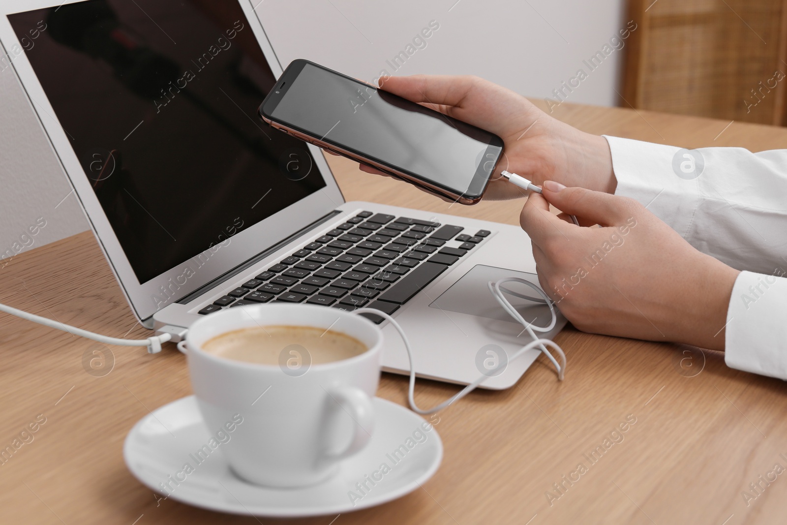 Photo of Woman connecting charging cable to smartphone near laptop at table indoors, closeup
